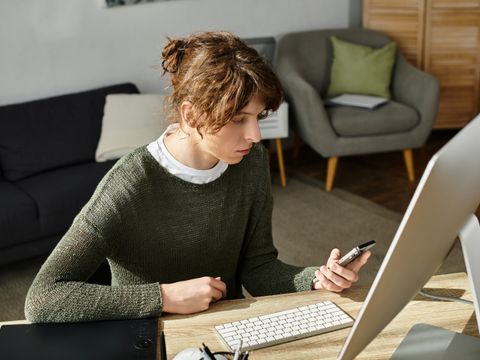 Frau mit lockigem Haar und grünem Pullover sitzt an einem Schreibtisch und schaut auf ihr Smartphone