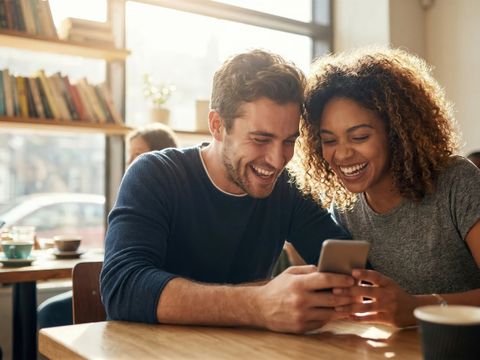 Zwei lächelnde Personen sitzen in einem Café und schauen gemeinsam auf ein Smartphone