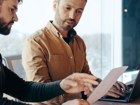 Zwei Männer sitzen an einem Tisch, einer zeigt dem anderen ein Blatt Papier, im Hintergrund ein Laptop