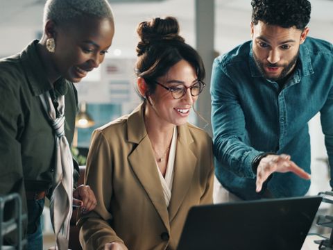 Drei Personen blicken lächelnd auf einen Laptop, eine Person zeigt mit der Hand darauf, alle stehen in einem modernen Büro