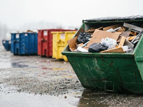 Ein großer grüner Industrie-Müllcontainer mit gemischte Bau- und Abbruchabfällen steht an einem nebligen Tag in einer Reihe bunter Abfallbehälter auf einem nassen Schotterplatz