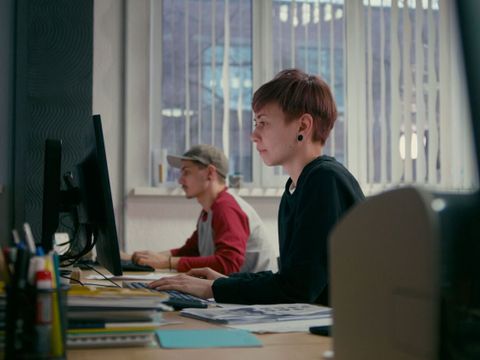 Zwei Personen sitzen an einem Schreibtisch vor Computermonitoren in einem Büro mit großen Fenstern