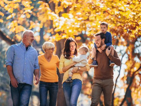 Fünf Personen einer Familie gehen im Herbstwald, zwei Erwachsene tragen Kleinkinder auf Armen und Schultern, alle lächeln
