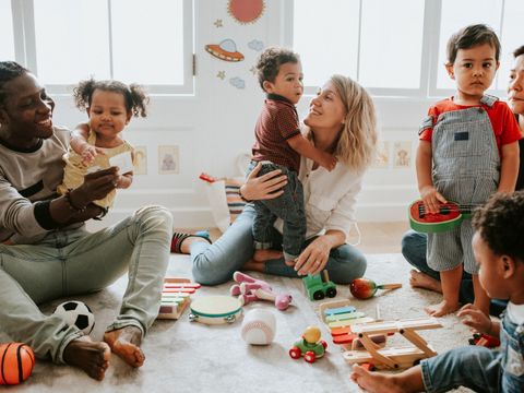 Mehrere Erwachsene und Kinder sitzen gemeinsam auf dem Boden eines hellen Raumes. Sie sind von buntem Holzspielzeug, einem Ball und Musikinstrumenten umgeben. Ein Kind sitzt auf dem Schoß einer Frau, während ein anderes Kind von einer weiteren Frau gehalten wird. Die Stimmung wirkt entspannt und freundlich. Im Hintergrund sind große Fenster und Wanddekorationen mit kindlichen Motiven zu sehen.