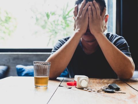Ein Mann schlägt die Hände vors Gesicht, vor ihm auf einem Tisch stehen ein Glas Alkohol, eine Dose Tabletten und ein Autoschlüssel