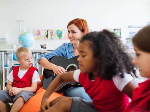 Schulkinder und Lehrerin mit Gitarre sitzen im Klassenzimmer, ein Kind mit geistiger Behindung