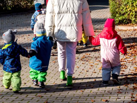 Eine erwachsene Person in heller Jacke hält die Hände von mehreren Kleinkindern, die bunte Winterkleidung tragen, beim Spaziergang auf einem gepflasterten Weg