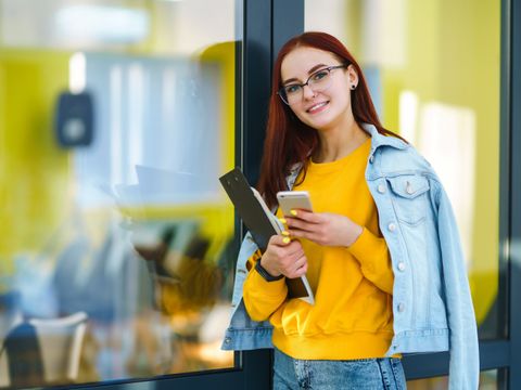 Frau mit gelbem Pullover und Jeansjacke lehnt an Glaswand und hält Smartphone und Ordner in der Hand