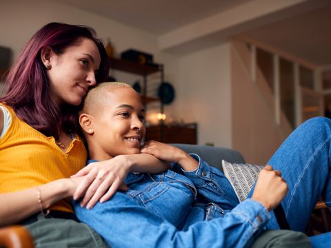 Zwei Frauen sitzen eng nebeneinander auf einem Sofa, eine lehnt sich an die andere und beide schauen lächelnd nach vorne
