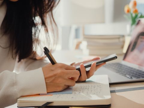 Person in weißem Pullover schreibt mit Stift in Notizbuch und hält Smartphone vor offenem Laptop auf hellem Schreibtisch