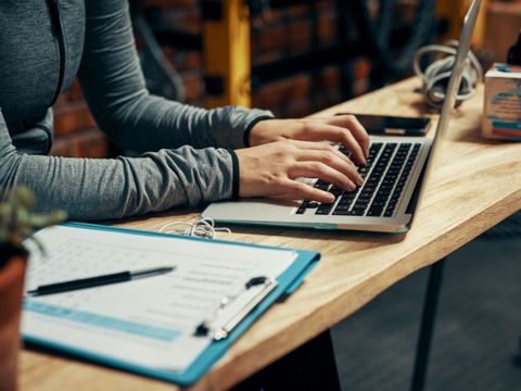 Person tippt auf Laptop an einem Holztisch. Neben dem Laptop liegen Klemmbrett und Stift.