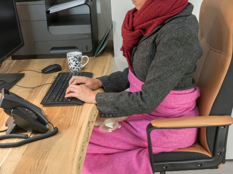 Frau sitzt am Schreibtisch im Büro und wärmt sich mit Decke, Schal und Wärmflasche