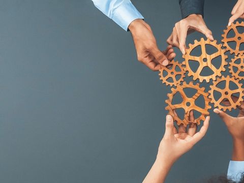 Mehrere Hände greifen nach ineinandergreifenden Zahnrädern aus Holz, symbolisieren Zusammenarbeit, Teamwork und das Zusammenspiel von Prozessen.