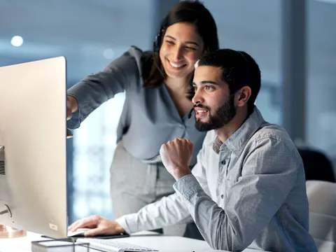 Ein Mann sitzt an einem PC-Arbeitsplatz und eine Frau steht neben ihm und zeigt ihm etwas auf dem Bildschirm