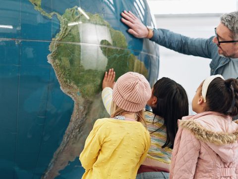 Lehrer und kleine Schülerinnen stehen an großem Globus und betrachten diesen