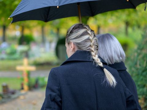 Zwei von hinten aufgenommene Personen mit dunklen Mänteln, eine Person hält einen Regenschirm, im Freien mit unscharfem Hintergrund aus Bäumen und Gräbern