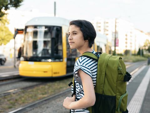 Person mit grünem Rucksack und gestreiftem T-Shirt steht an einer Straßenbahnhaltestelle vor einer gelben Straßenbahn in Berlin
