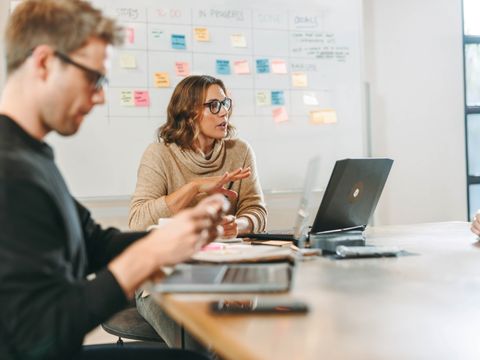 Frau mit Brille spricht an einem Tisch mit Laptop vor einer Wand mit bunten Notizzetteln, Mann im Vordergrund hat vor sich einen Laptop