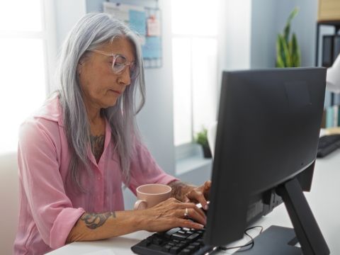Frau mit grauen Haaren und Brille sitzt an einem Schreibtisch und tippt auf einer Tastatur vor einem großen Monitor