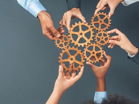 Mehrere Hände greifen nach ineinandergreifenden Zahnrädern aus Holz, symbolisieren Zusammenarbeit, Teamwork und das Zusammenspiel von Prozessen.