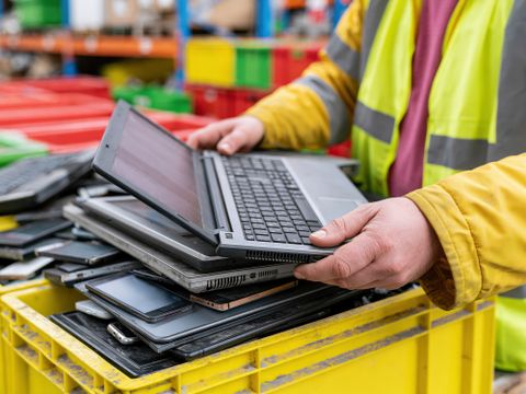 Ein Arbeiter in einer gelben Sicherheitsweste sortiert in einem Lagerhaus alte Laptops und Handys für das Recycling