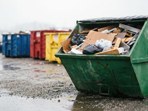 Ein großer grüner Industrie-Müllcontainer mit gemischte Bau- und Abbruchabfällen steht an einem nebligen Tag in einer Reihe bunter Abfallbehälter auf einem nassen Schotterplatz