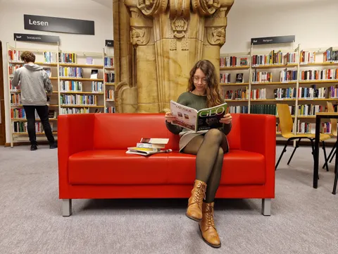 Frau mit Zeitschrift auf einem roten Sofa vor Bibliotheksregalen