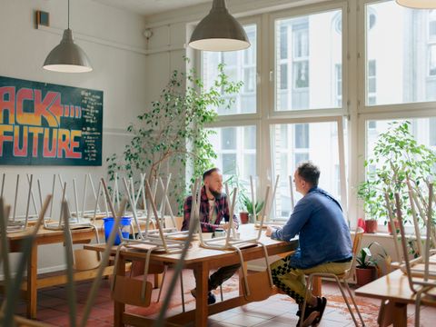 Die stellvertretende Leitung der Übergangseinrichtung, Stephan Boer, Erzieher und Arbeitstherapeut, sitzt an einem Tisch und führt ein Gespräch mit einem Klienten. Im Raum stehen mehrere Tische und Stühle, die Stühle sind auf den Tischen gestapelt. Durch die großen Fenster fällt viel Tageslicht, und grüne Pflanzen schmücken den Raum. An der Wand hängt ein „Back to the Future“-Poster.