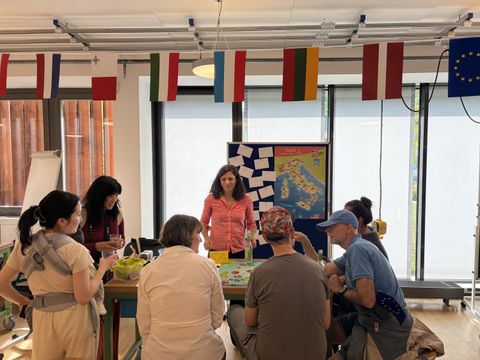 Bildbeschreibung: Eine Sprachlehrerin mit sechs Menschen an ihrem Tisch, die im Gespräch sind. Im Hintergrund eine Aufsteller mit Notizen und einer Italienkarte.