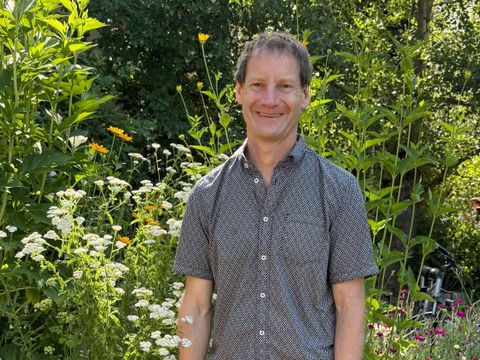 Ein Mann mit kurzem, braunem Haar steht vor hohen, blühenden Pflanzen in einem Garten. Er trägt ein kurzärmeliges, dunkelgraues Hemd mit feinem Muster und lächelt leicht. Im Hintergrund sind grüne Blätter, weiße und gelbe Blüten sowie ein Teil eines hellen Gebäudes zu sehen. Die Szene wirkt sommerlich und freundlich.