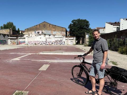 Alexander Matthes steht auf einer gepflasterten, offenen Fläche und hält ein schwarzes Fahrrad am Lenker. Er trägt ein kurzärmliges, kariertes Hemd und eine kurze Hose. Im Hintergrund sind eine hohe Backsteinmauer, mehrere mit Graffiti besprühte Wände und einige Bäume zu sehen. Der Himmel ist wolkenlos und blau. Die Szene wirkt urban und sommerlich.