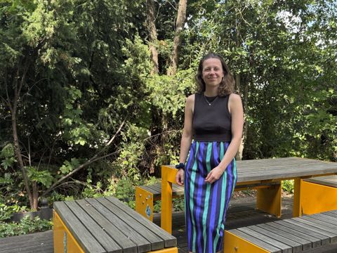 Eine Frau mit lockigem, schulterlangem Haar steht neben gelben Holzbänken und -tischen im Freien. Sie trägt ein ärmelloses schwarzes Oberteil und einen langen Rock mit vertikalen Streifen in Blau, Grün und Lila. Im Hintergrund sind dichte, grüne Bäume und Sträucher zu sehen. Die Szene wirkt sommerlich und freundlich.