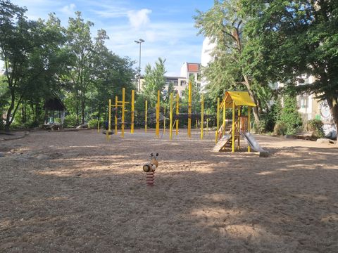 Im Mittelpunkt steht ein weitläufiger Sandspielplatz in der Körtestraße, umgeben von hohen Bäumen und angrenzenden Wohnhäusern. Zentral verlaufen mehrere gelbe Kletterstangen in parallelen Reihen. Rechts befindet sich ein Klettergerüst mit Rutsche und überdachtem Podest, ebenfalls in Gelb gehalten. Im Vordergrund steht eine Wippe in Tierform auf einer Spiralfeder. Links am Rand ist ein überdachter Pavillon mit Sitzgelegenheit zu erkennen. Der Himmel ist blau mit wenigen Wolken, das Gelände wirkt gepflegt und offen.