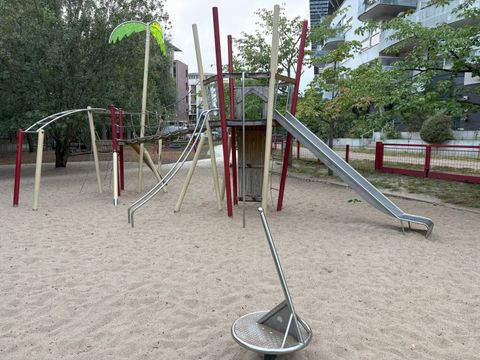 Im Vordergrund befindet sich eine moderne Spielplatzanlage mit hellen Sandflächen. Die Spielgeräte bestehen aus Metall und Holz, darunter eine lange Rutsche, eine Kletterstruktur mit schrägen und gebogenen Stangen sowie eine kleine drehbare Sitzfläche. Ein hölzernes Spielhaus mit roten und beigen Stützen steht zentral, darüber ragt eine stilisierte Palme aus Holz mit grünen Blättern. Im Hintergrund sind Bäume und mehrstöckige Wohnhäuser mit Balkonen zu sehen. Der Spielplatz ist von einem roten Zaun umgeben.