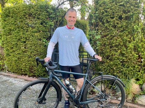 Eine Person steht auf einer gepflasterten Fläche vor einer dichten, grünen Hecke und hält ein modernes Rennrad mit dunklem Rahmen. Die Person trägt ein helles T-Shirt, eine kurze Hose, Sportschuhe und eine Sportuhr. Um den Hals hängt ein rotes Band mit einer Medaille. Die Hände ruhen auf dem Lenker des Fahrrads. Im Hintergrund sind weitere Pflanzen und Bäume zu sehen.