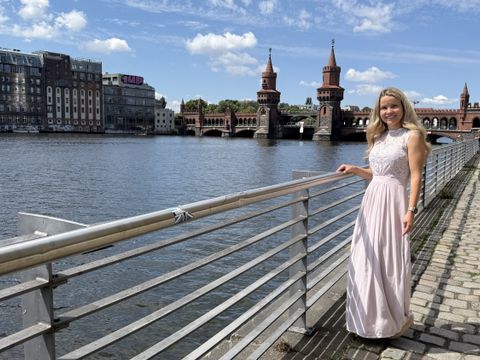 Eine Frau mit langen, blonden Haaren steht an einem Geländer am Ufer eines Flusses. Sie trägt ein helles, ärmelloses Kleid mit Spitzenoberteil und lächelt in die Kamera. Im Hintergrund ist die Oberbaumbrücke mit ihren markanten Türmen und einem gelben Zug zu sehen. Der Himmel ist blau mit wenigen Wolken.