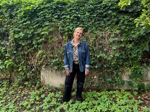 Eine Person steht vor einer dichten, grünen Efeuwand. Sie trägt eine blaue Jeansjacke, ein gemustertes Oberteil und eine schwarze Hose. Der Boden ist mit niedrigen Pflanzen bedeckt. Die Person blickt in die Kamera und lächelt leicht.