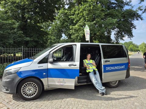Ein blau-weißer Kleintransporter mit der Aufschrift 'ORDNUNGSAMT' steht am Straßenrand. Die Schiebetür auf der Beifahrerseite ist geöffnet. Eine Person mit gelber Warnweste sitzt entspannt auf dem Fahrzeugsitz, lehnt sich zurück und hat die Beine ausgestreckt. Im Hintergrund sind grüne Bäume und ein Metallzaun zu sehen. Das Wetter wirkt sonnig und freundlich.