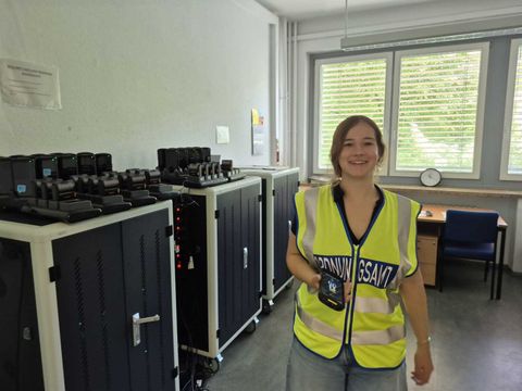 Eine Frau mit langen, offenen Haaren steht in einem Raum mit mehreren großen, schwarzen Geräteschränken an der Wand. Sie trägt eine gelb-leuchtende Warnweste mit reflektierenden Streifen und hält ein kleines elektronisches Gerät in der rechten Hand. Im Hintergrund befinden sich ein Schreibtisch mit einem Stuhl, eine Wanduhr und ein Fenster mit Jalousien. Auf den Geräteschränken sind zahlreiche identische, aufrecht stehende Geräte zu sehen.