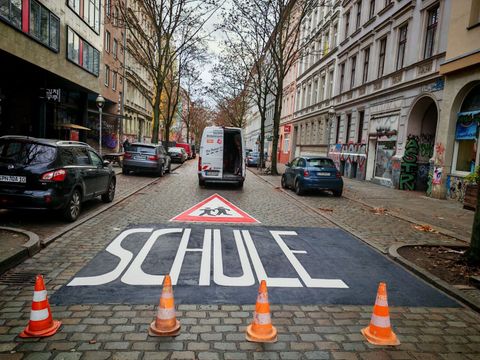 Im Vordergrund ist eine großflächige Straßenmarkierung auf dem Kopfsteinpflaster der Manteuffelstraße zu sehen. Die Markierung besteht aus einem weißen Dreieck mit rotem Rand und einem Piktogramm zweier gehender Kinder, das auf einen Schulweg hinweist. Darunter steht in großen weißen Buchstaben das Wort 'SCHULE'. Drei orangefarbene Leitkegel grenzen den Bereich ab. Links und rechts der Straße parken Fahrzeuge, im Hintergrund steht ein Lieferwagen. Die Szene wird von mehrstöckigen Altbauten und kahlen Bäumen gesäumt. Die Markierung betont die Schulwegsicherheit an dieser Stelle.