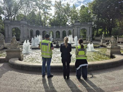 Drei Personen stehen mit dem Rücken zur Kamera vor einem großen, runden Brunnen mit mehreren Wasserfontänen. Sie tragen gelbe Warnwesten mit reflektierenden Streifen und dunkle Kleidung. Im Hintergrund ist eine steinerne Balustrade mit Bögen und Skulpturen zu sehen, dahinter hohe Bäume. Die Szene spielt sich bei Tageslicht ab, der Himmel ist teilweise bewölkt.