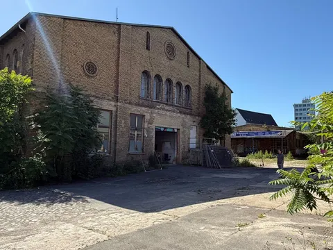 Backsteingebäude mit hohen Rundbogenfenstern und dekorativen Zierelementen an der Fassade, das als Alte Reithalle auf dem Dragonerareal bekannt ist. Im Vordergrund ein asphaltierter Hof mit vereinzeltem Bewuchs und Schattenwurf. Links und rechts wachsen größere Sträucher und Bäume an der Gebäudeseite. Im Hintergrund sind weitere Gebäude und ein moderneres Hochhaus zu sehen. Der Himmel ist wolkenlos und blau.