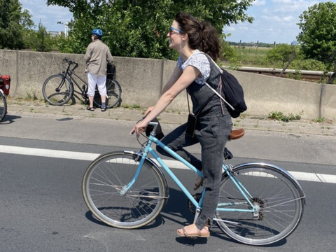 Eine Frau mit lockigen, dunklen Haaren fährt auf einem hellblauen Fahrrad auf einer asphaltierten Straße. Sie trägt einen schwarzen Rucksack, ein graues Oberteil und eine graue Hose. Im Hintergrund stehen zwei weitere Personen mit Fahrrädern an einer niedrigen Betonmauer, eine davon trägt einen blauen Helm und eine rote Fahrradtasche. Bäume und ein blauer Himmel mit wenigen Wolken sind sichtbar.