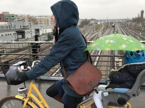 Sara Lühmann fährt auf einem gelben Fahrrad über eine Brücke. Sie trägt eine blaue Jacke mit Kapuze, eine braune Umhängetasche und braune Stiefel. Auf dem hinteren Kindersitz sitzt ein Kind mit blauer Jacke, weißer Hose und pinken Schuhen. Das Kind hält einen grünen Regenschirm mit weißen Punkten. Im Hintergrund sind Bahngleise und städtische Gebäude zu sehen. Die Szene spielt im Rahmen von Stadtradeln 2026.