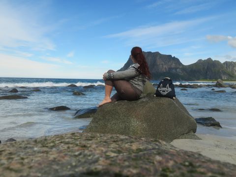 Eine Person mit langen, rötlichen Haaren sitzt barfuß auf einem großen Stein am Strand und blickt aufs Meer. Sie trägt eine graue Strickjacke mit Muster und braune Hose. Neben ihr steht ein dunkler Rucksack mit einem weißen Motiv. Im Hintergrund sind Wellen, ein heller Sandstreifen und eine markante, grüne Bergkette unter blauem Himmel zu sehen.