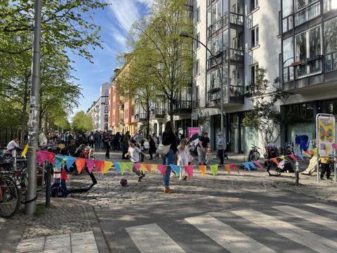 Im Vordergrund verläuft eine bunte Wimpelkette quer über die gepflasterte Straße, die als Schulzone Simplon-/Niemannstraße gekennzeichnet ist. Kinder spielen mit einem Ball auf der Straße, während Erwachsene am Rand stehen oder sitzen. Links sind Fahrräder abgestellt, rechts steht ein Stadtplan mit farbigen Markierungen. Die Umgebung ist von mehrgeschossigen Wohnhäusern und grünen Bäumen geprägt. Die Szene vermittelt eine lebendige, gemeinschaftliche Atmosphäre.