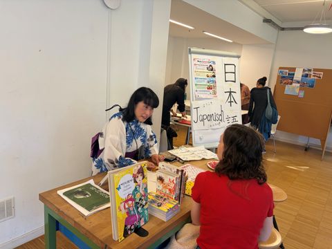 Eine Frau im Gespräch mit einer anderen Person an einem Stand mit japanischen Büchern. Im Hintergrund ein Flipchart mit japanischen Schriftzeichen.