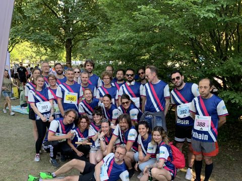 Eine größere Gruppe von Menschen steht und sitzt im Freien auf einer Wiese unter Bäumen. Alle tragen einheitliche Sporttrikots mit diagonalen Streifen in Blau, Weiß und Rot sowie Startnummern. Einige Personen halten Getränkeflaschen in der Hand. Im Hintergrund sind weitere Menschen und Bäume zu sehen. Die Szene wirkt entspannt und gemeinschaftlich, typisch für ein Teamfoto nach einer Sportveranstaltung wie der Teamstaffel 2024.