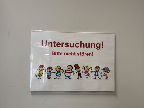 Ein rechteckiges Schild mit weißem Hintergrund zeigt oben in großer, roter Schrift den Hinweis 'Untersuchung!'. Darunter steht in kleinerer, ebenfalls roter Schrift: 'Bitte nicht stören!'. Am unteren Rand ist eine bunte Zeichnung von sieben Kindern zu sehen, die nebeneinander stehen und unterschiedliche Kleidung tragen.