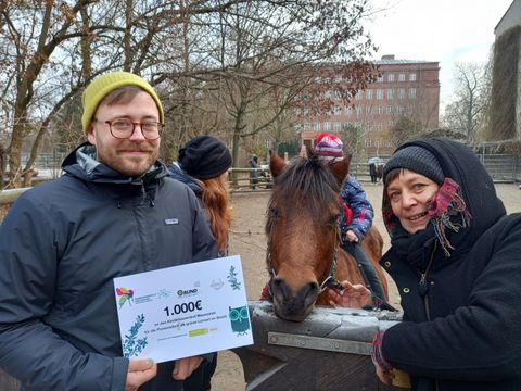 Im Vordergrund zwei Personen, eine davon hält Spendencheck in der Hand mit Aufschrift 1.000 Euro. Im HIntergrud ist ein Kind auf einem Pony.